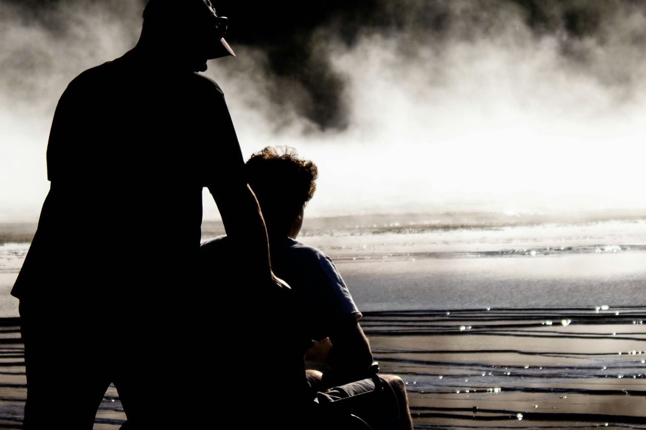 Une personne accompagne un proche en fauteuil roulant au bord d'un plan d'eau, à contre-jour.