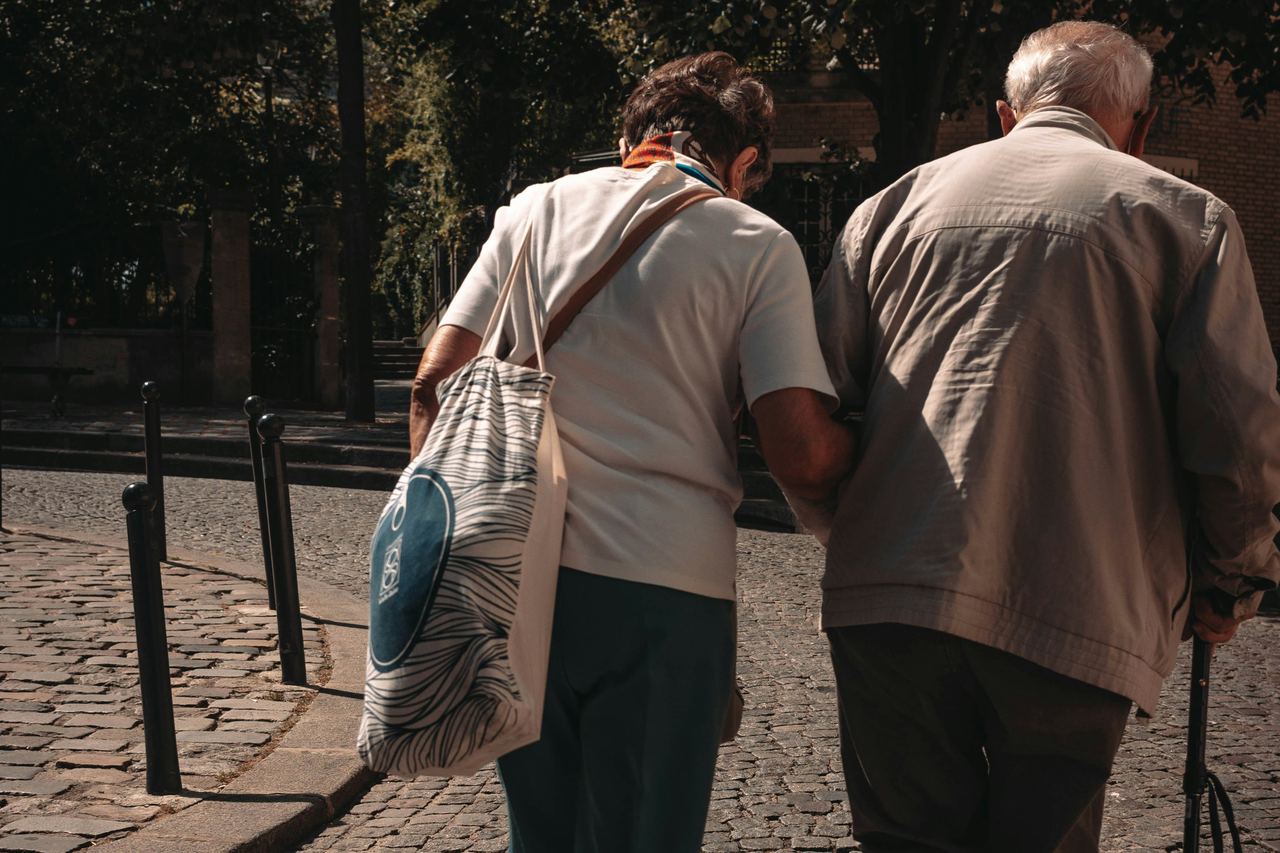 Une femme soutient par le bras un homme âgé qui marche avec une canne, sur une rue pavée.